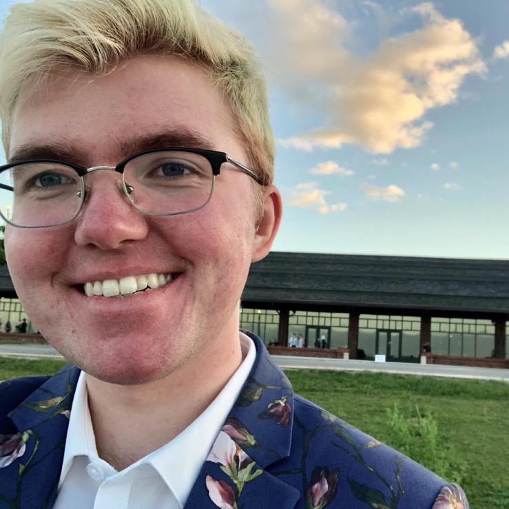 Leland Ashford Lanquist smiling, in a floral dark blue suit and a crisp white shirt, with square glasses, bleached hair, and pink skin. In the background is a lawn in front of a long building and a pretty sky with a few sunlit clouds.